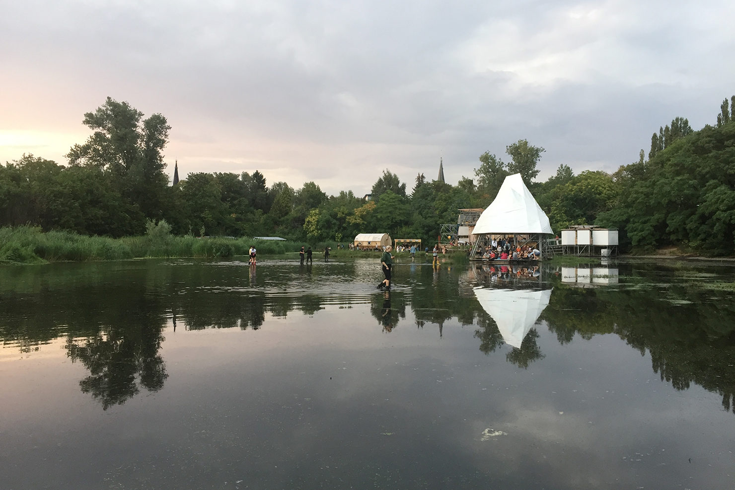 A photo of the Floating University Berlin. It is a collection of small wooden structures supported by legs which raise it above a concrete basin, which is captured here, flooded, after recent rainfall. The sun the going down. There are trees and church spires in the background. A blanket of light clouds fills the sky. 