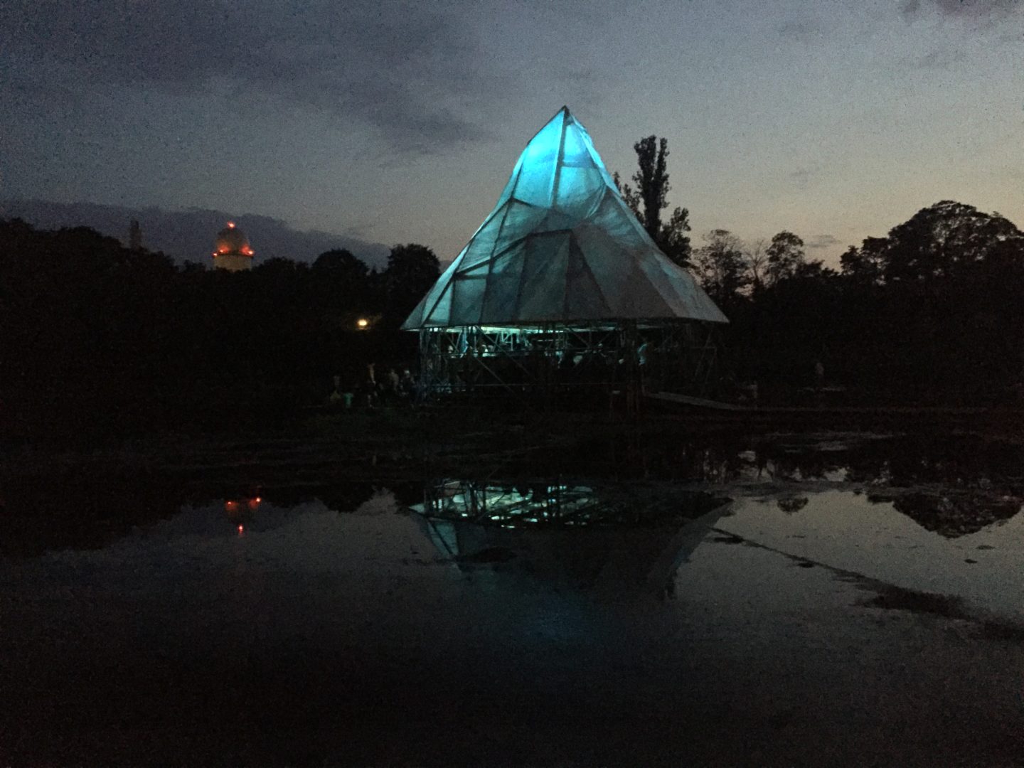 The Floating University pictured at night. The depicted structure is illuminated light blue and resembles an iceberg. It is surrounded by darkness. Water in the foreground reflects the light.