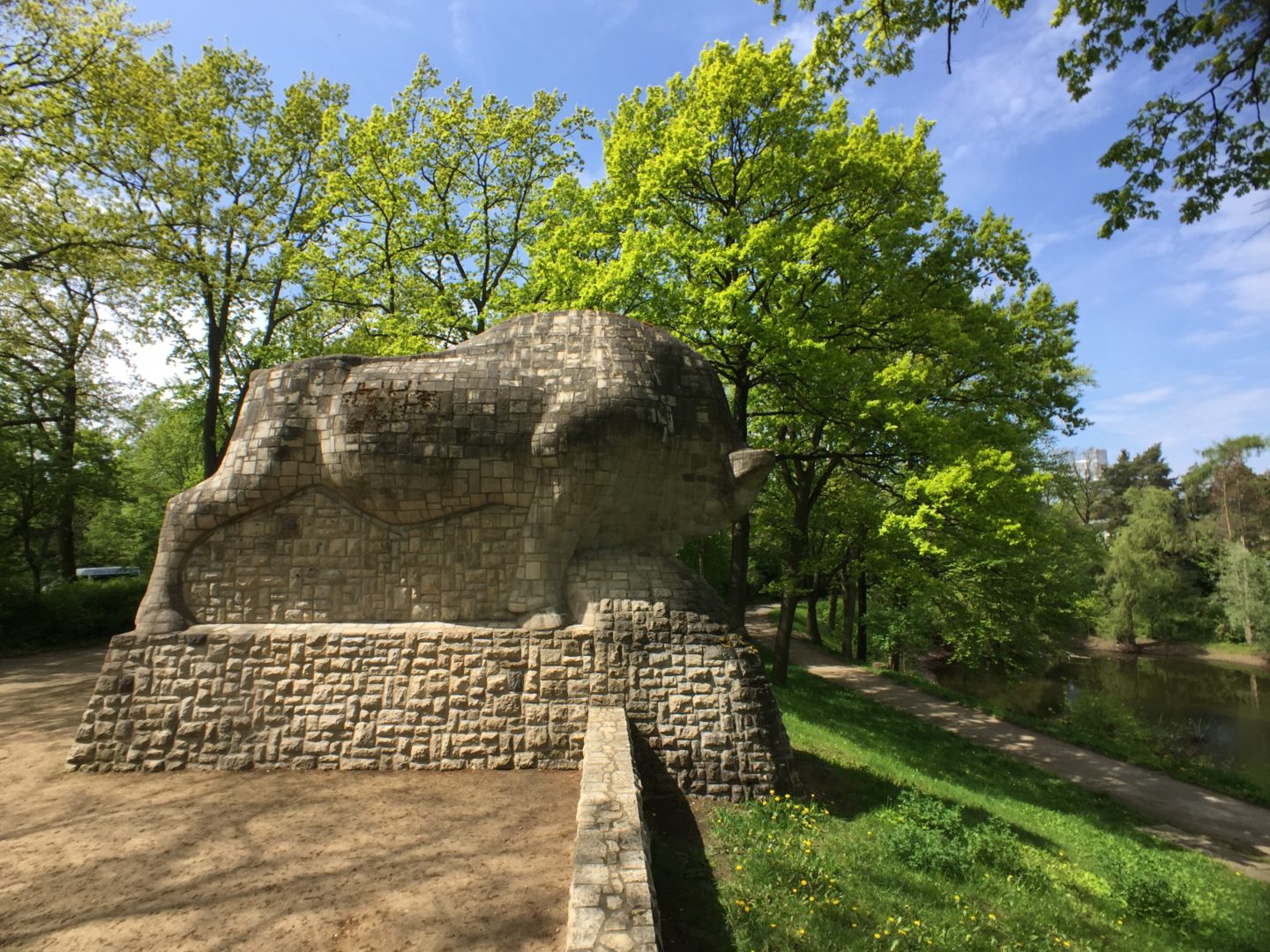 Paul Mersmann's 1934 sculpture of an aurochs overlooking the pond Blanke Helle. The sculpture is very large, much higher than an adult, and made entirely of carved stone. It was photographed in sunlight. The sky is blue. It is surrounded by the greenery of big trees.